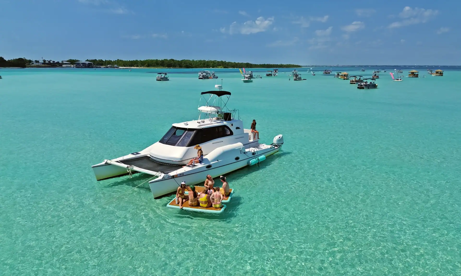 A white yacht with guests relaxing on Crab Island's turquoise waters, surrounded by numerous boats and a vibrant summer scene.