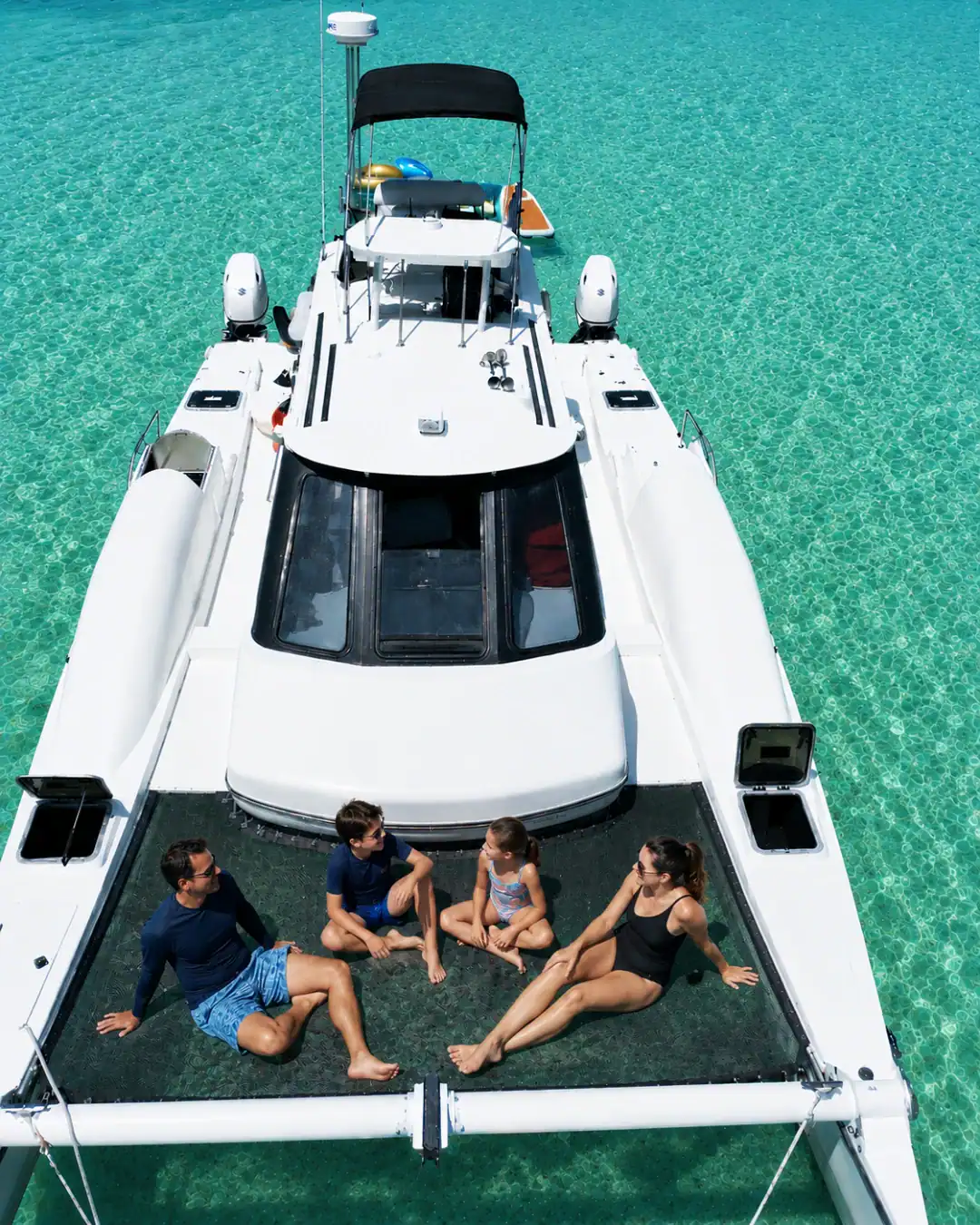 Family, sitting on the net of a catamaran anchored at Crab Island in crystal clear emerald water.